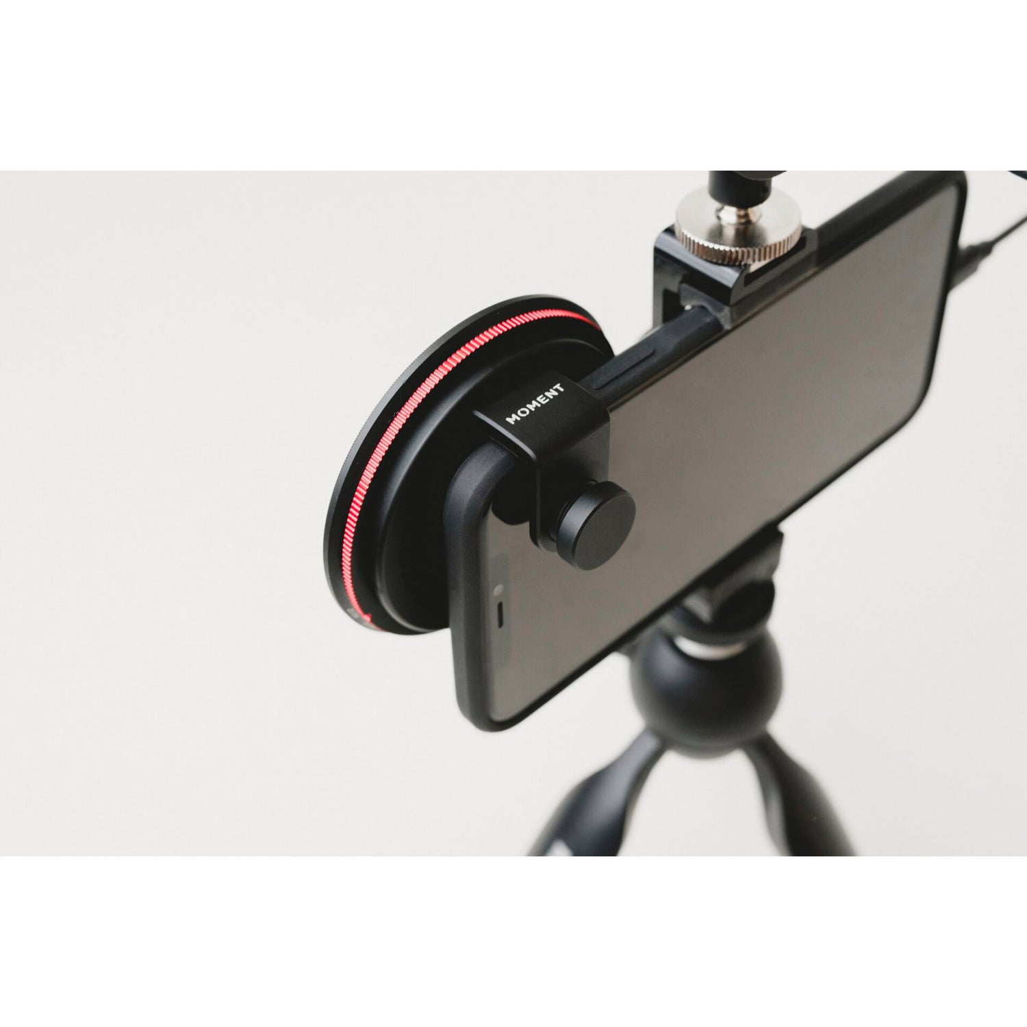 Ein Smartphone auf einem Stativ wird mit dem MOMENT 67 mm Universal Phone Case kompatibler Filter Mount mit praktischem Filteradapter vor einem schlichten Hintergrund gezeigt.