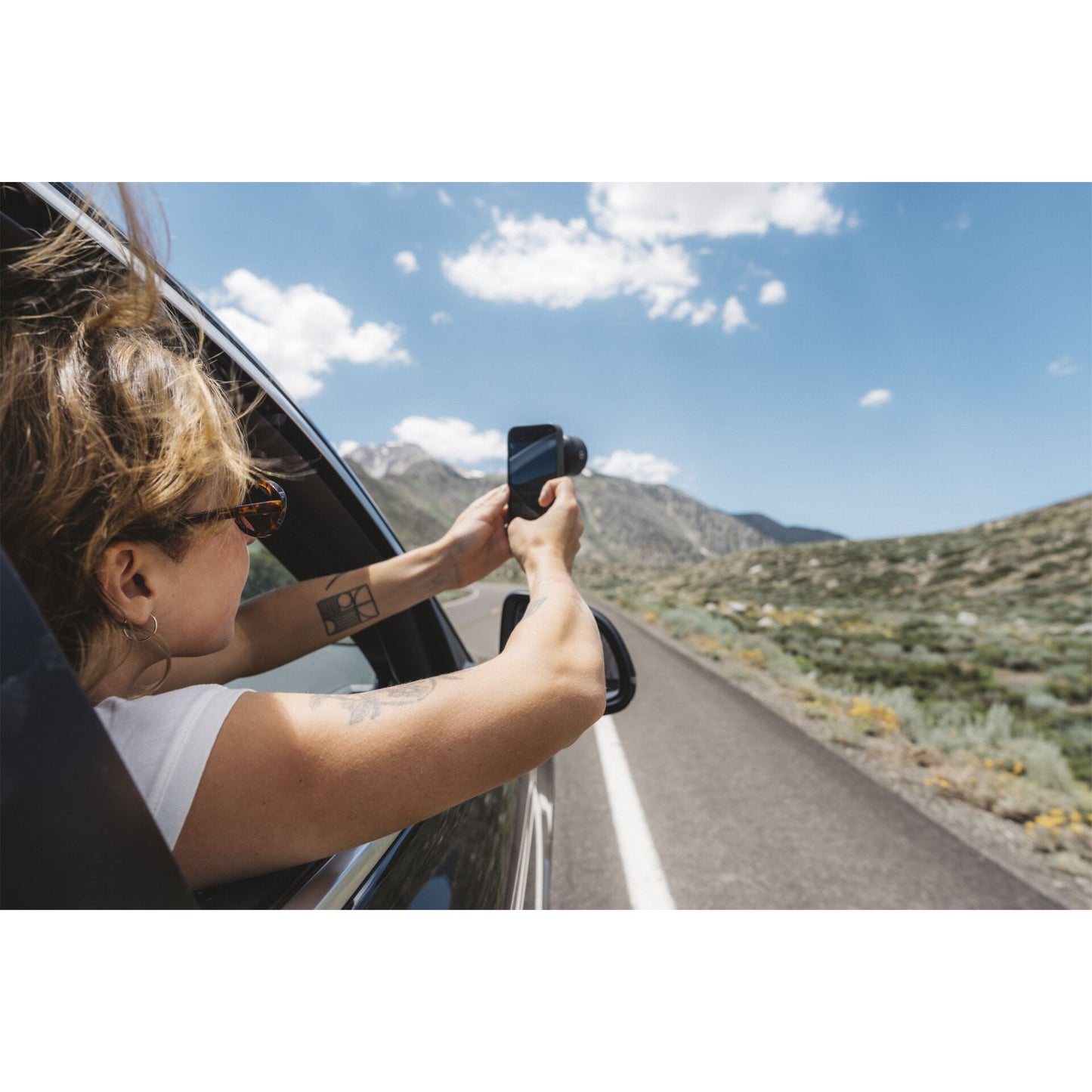 Eine Person lehnt aus dem Autofenster und fotografiert die malerische Bergstraße bei Sonnenschein mit dem MOMENT 58mm Tele Lens T-Series für optische Vergrößerung.