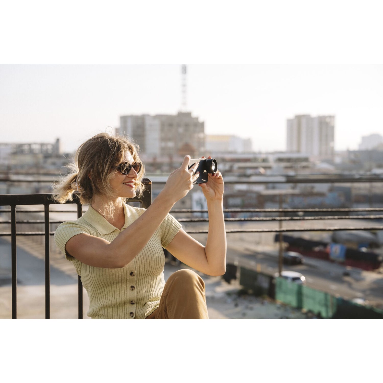 Eine Frau mit Sonnenbrille macht ein Dachfoto von der Stadt und verwendet dabei ihr MOMENT 58-mm-Teleobjektiv der T-Serie für einen beeindruckenden optischen Zoom.