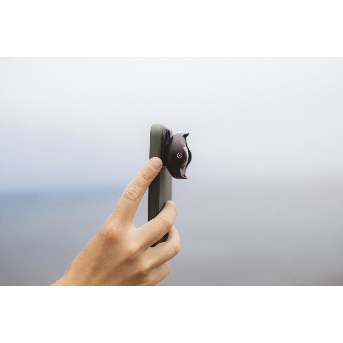 Eine Hand hält ein iPhone mit dem MOMENT 18-mm-Weitwinkelobjektiv - T-Serie, einer Premium-Kameraausrüstung, vor einem unscharfen Hintergrund im Freien.