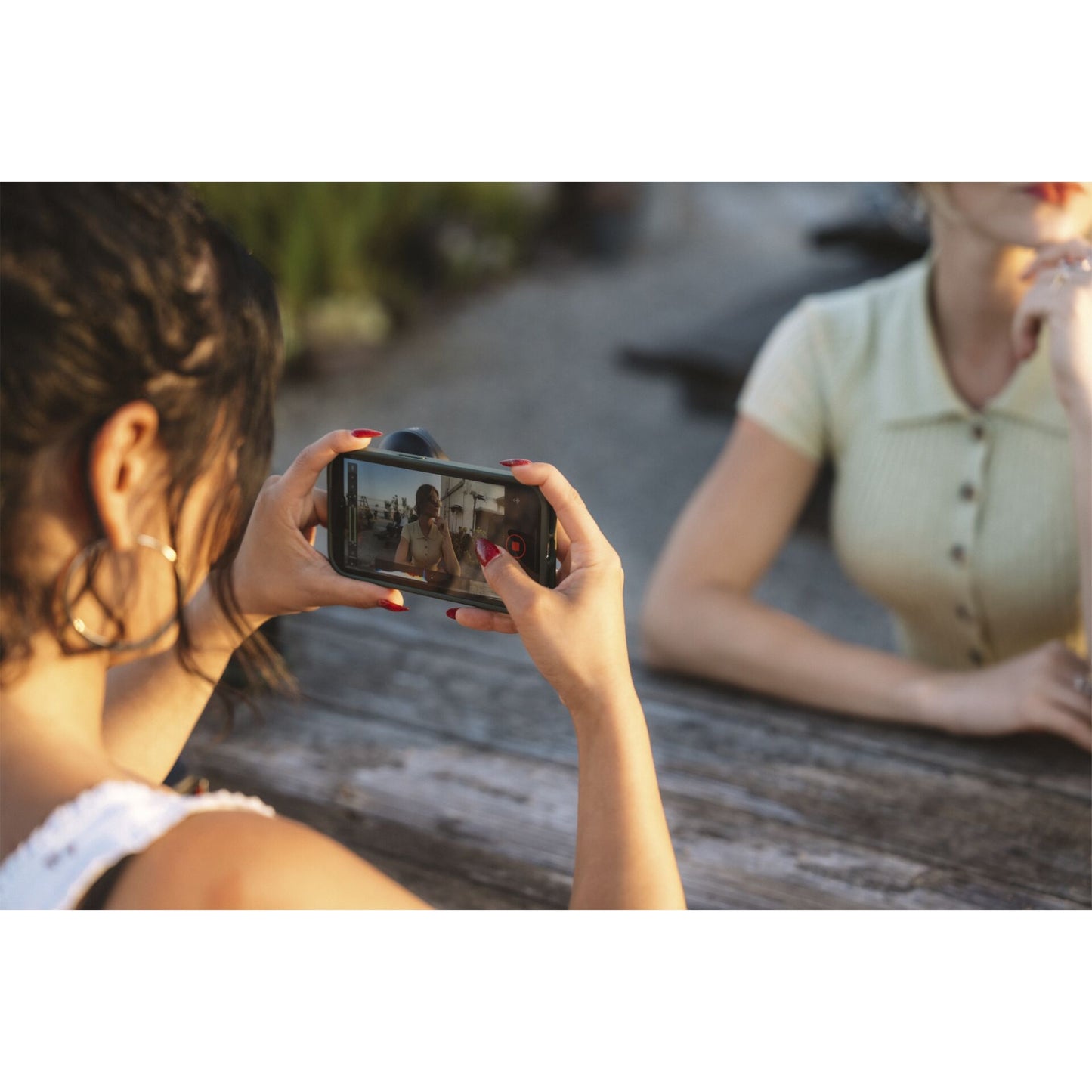 Eine Person verwendet das MOMENT Moment 18mm Weitwinkelobjektiv - T-Serie, um mit ihrem Smartphone ein Foto von einem Freund zu machen, der im Freien an einem Holztisch sitzt.