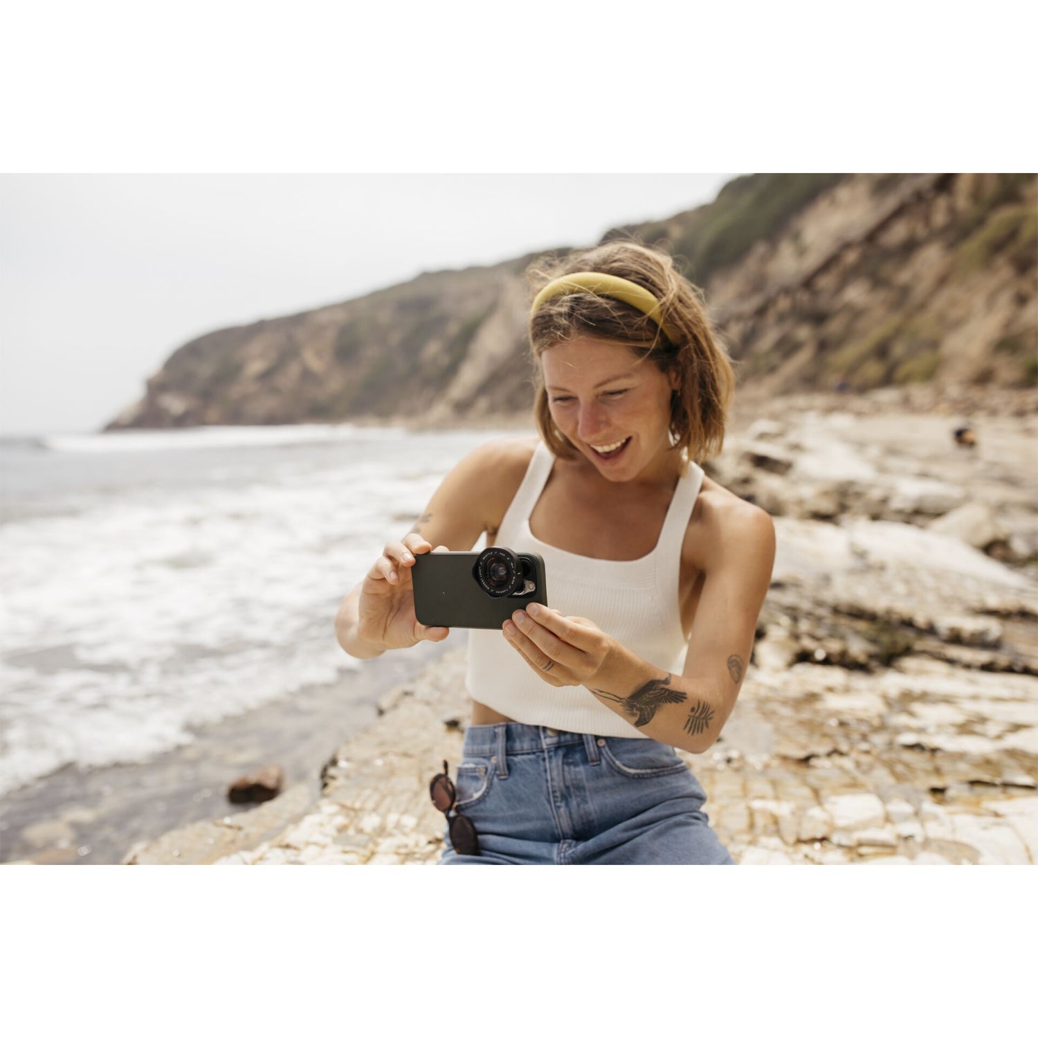 Eine lächelnde Frau macht mit dem MOMENT 18-mm-Weitwinkelobjektiv - T-Serie ein Foto mit ihrem Handy an einem felsigen Strand, mit dem Meer und den Klippen im Hintergrund.