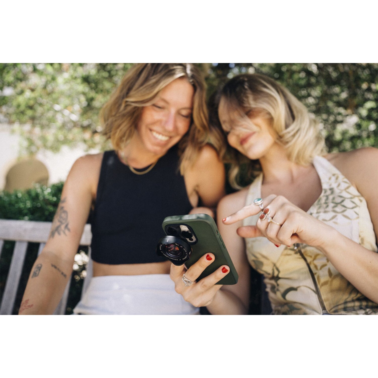 Zwei Frauen sitzen auf einer Bank und blicken lächelnd auf ein Smartphone - Erinnerungen festhalten mit dem MOMENT 18mm Weitwinkelobjektiv - T-Serie, dem idealen Zubehör für atemberaubende iPhone Outdoor-Fotos.