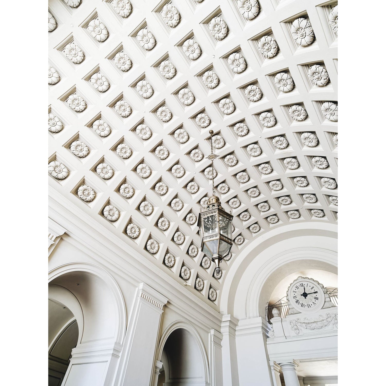 Dekorative weiße Gewölbedecke mit einer hängenden Laterne und einer großen Uhr über gewölbten Türöffnungen, wunderschön eingefangen mit dem MOMENT 18mm Weitwinkelobjektiv - T-Serie für Ihr iPhone-Kamerazubehör.