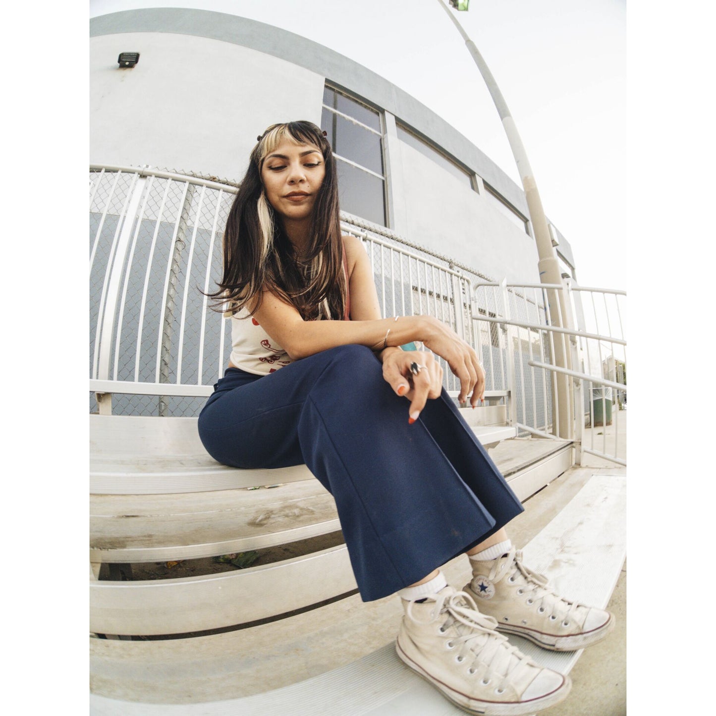 Frau mit langen Haaren und Pony sitzt in marineblauer Hose und weißen Turnschuhen auf einer Tribüne im Freien. Perfekt eingefangen mit einem iPhone-Kamerazubehör und dem MOMENT 14mm Fisheye-Objektiv - T-Series.
