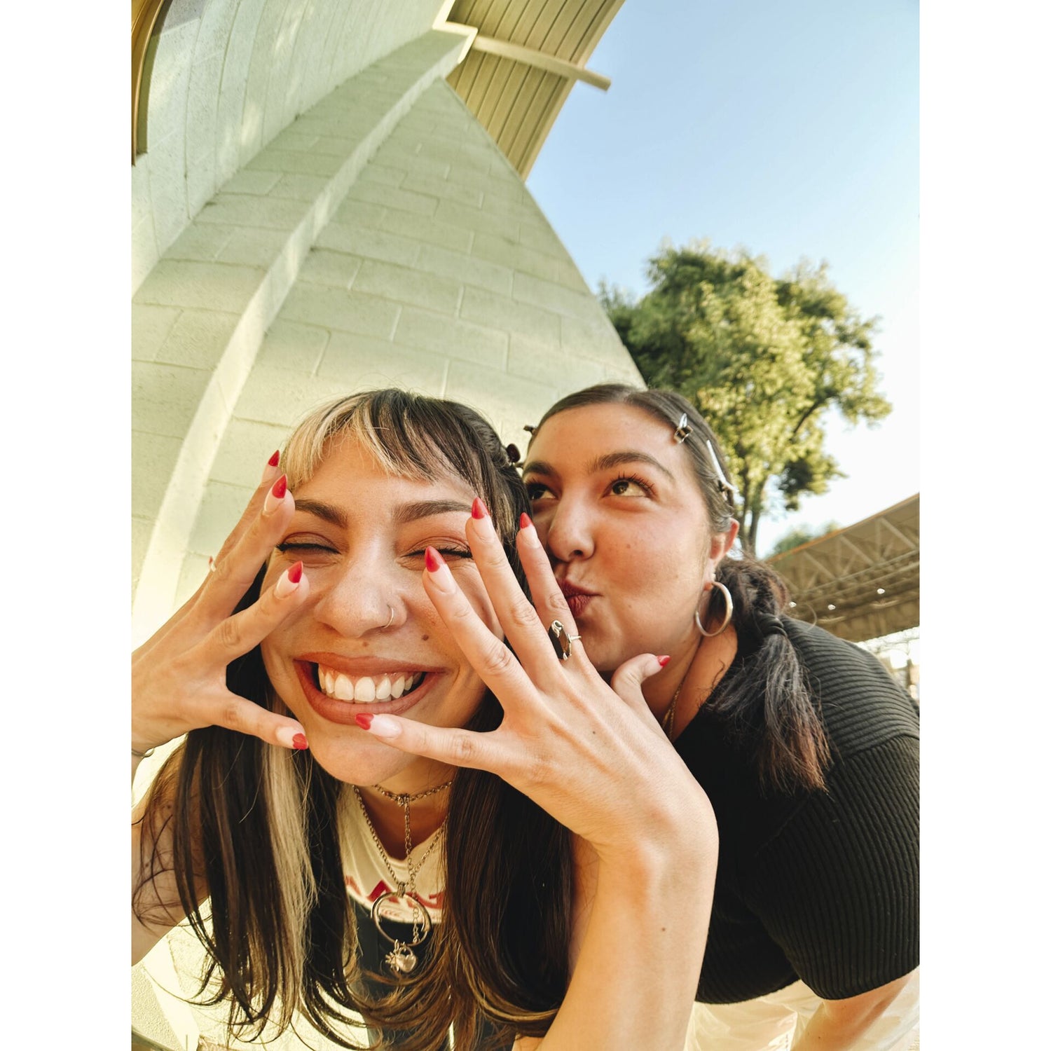 Zwei Freundinnen posieren spielerisch im Freien; die eine lächelt mit den Händen im Gesicht, während die andere ihre Wange küsst. Eingefangen wurde der freudige Moment mit einem MOMENT 14mm Fisheye-Objektiv - T-Serie.