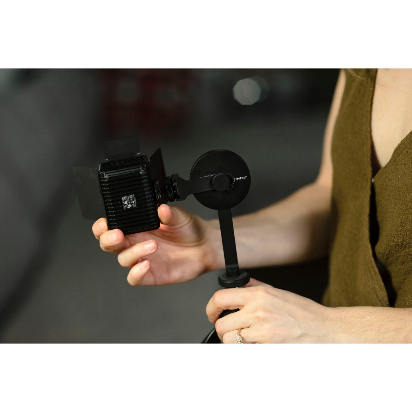 Person mit ärmellosem Oberteil, die mit dem MOMENT Moment Pro Tripod Mount für MagSafe und einem iPhone eine kleine Kameralampe auf einem Handstativ ausrichtet.