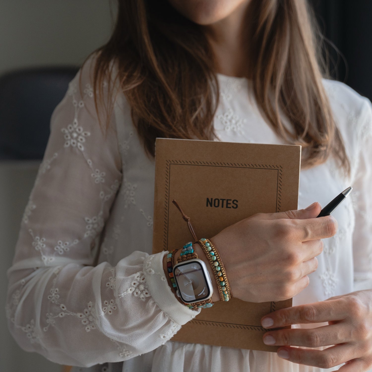 Eine Frau in einem weißen Spitzenkleid hält ein Notizbuch und einen Stift in der Hand und trägt dazu bunte Armbänder, eine Uhr und das arktisband Apple Watch Armband "Dream Catcher" von arktisband für einen schicken Touch.