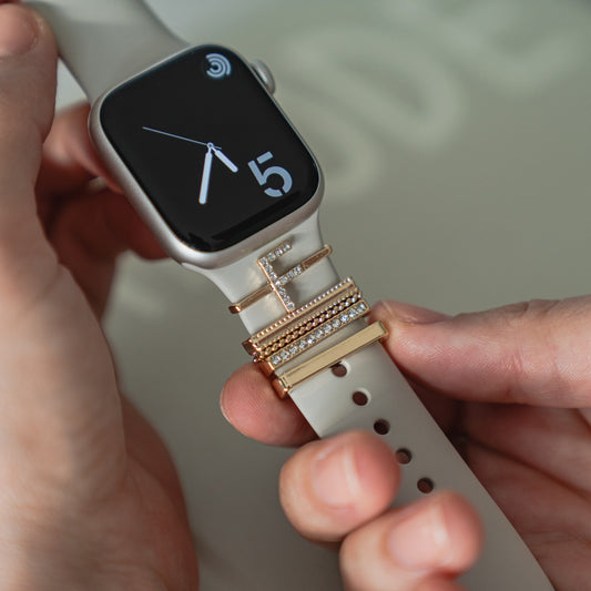 Eine Hand hält eine Smartwatch mit mehreren goldenen und diamantenen Arktisband Apple Watch Charms „F Style“ von Arktisband, die auf dem weißen Armband gestapelt sind und so für einen luxuriösen Look sorgen.