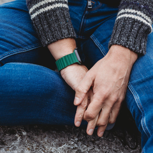 Eine Person trägt das arktisband Apple Watch Magnetarmband „Neo“ von arktisband und einen dunklen, gestreiften Pullover. Sie sitzt im Schneidersitz und stützt ihre Hände auf die Knie. Nur der Unterkörper, die Arme und die Hände sind sichtbar.