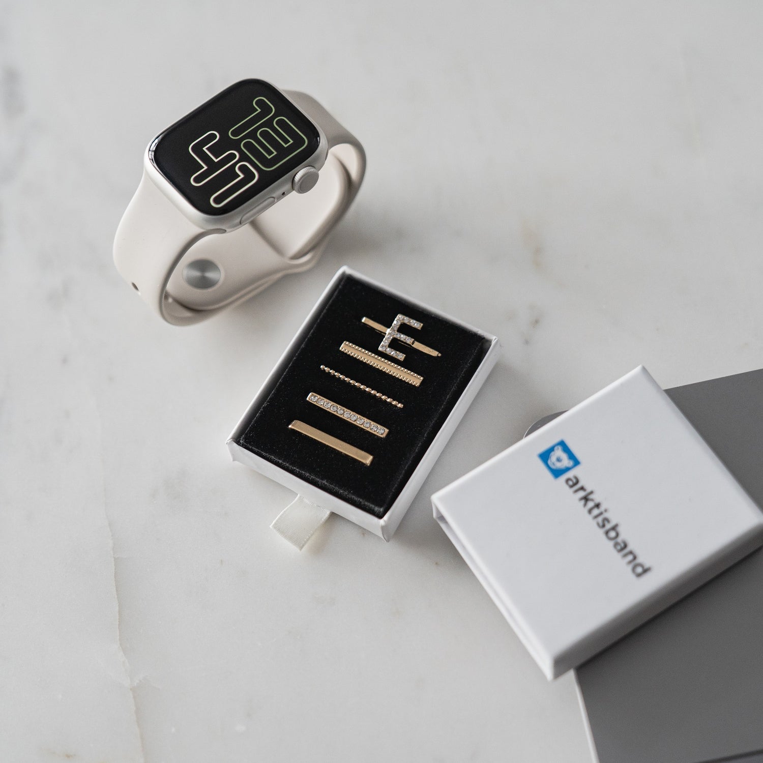Ein arktisband Apple Watch Charms „E Style“-Set – stylische Metallarmbänder von arktisband – liegt auf einer Marmoroberfläche neben einer Smartwatch und einer offenen Box.