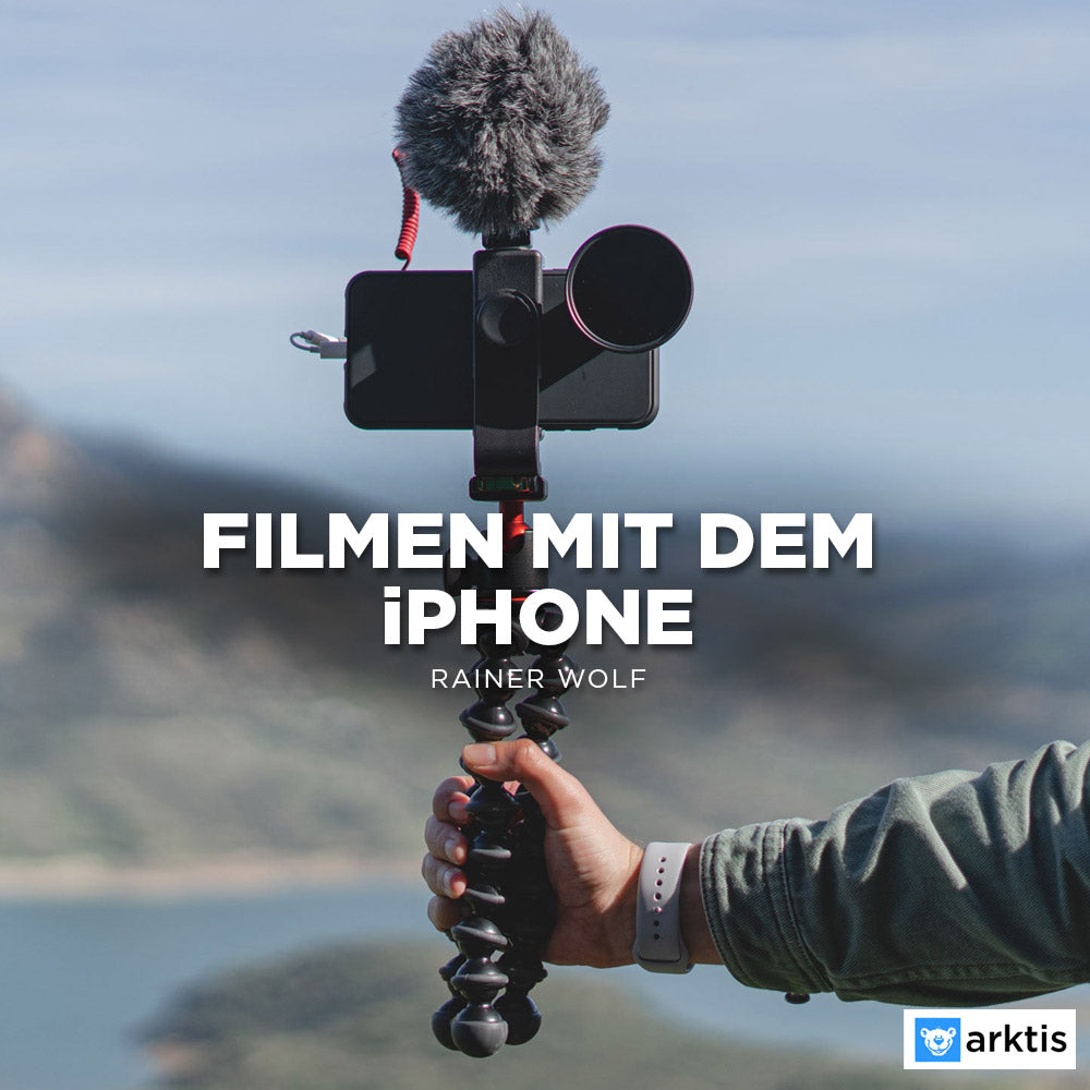 Eine Person hält ein flexibles, auf einem Stativ montiertes iPhone mit Mikrofon und Objektiv im Freien mit verschwommenen Hügeln und Wasser. Text: Filmen mit dem iPhone – Online-Kurs von Rainer Wolf.