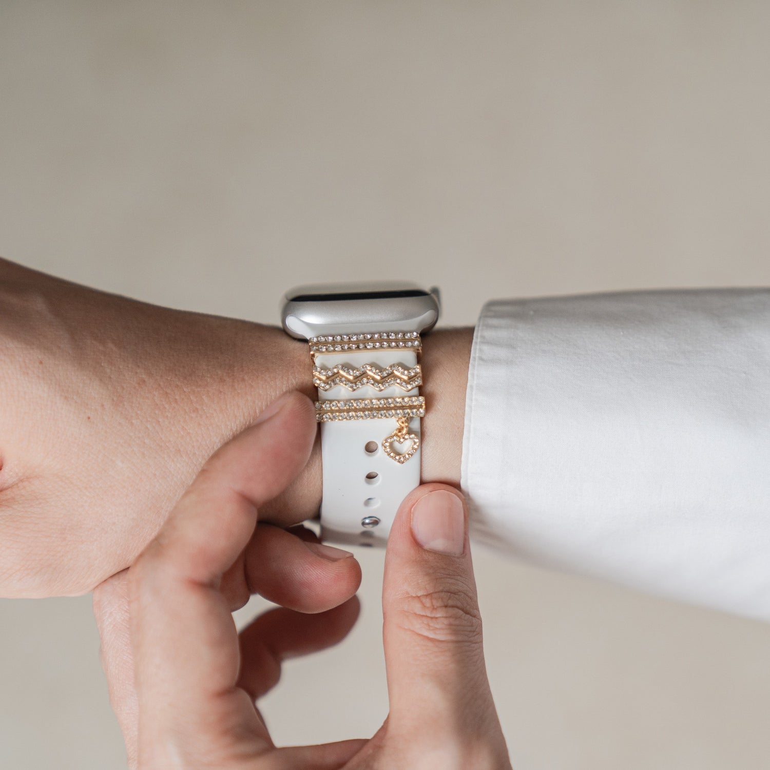 Eine Person trägt dekorative Goldarmbänder und die Arktisband Apple Watch Charms „Hanging Heart“ – Gold am Handgelenk und rückt das Armband ihrer Smartwatch zurecht.