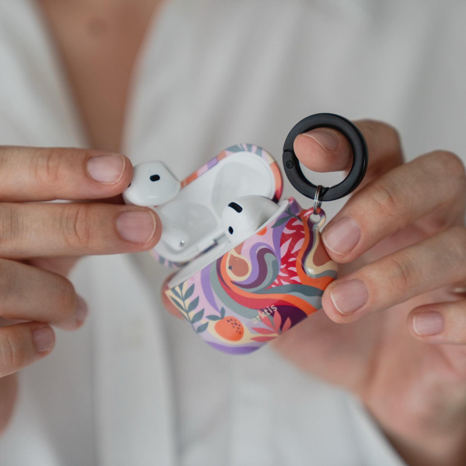 Hände, die eine geöffnete Arktis AirPods 5-Hülle halten – Summer mit Blumenmuster und schwarzem Befestigungsring.