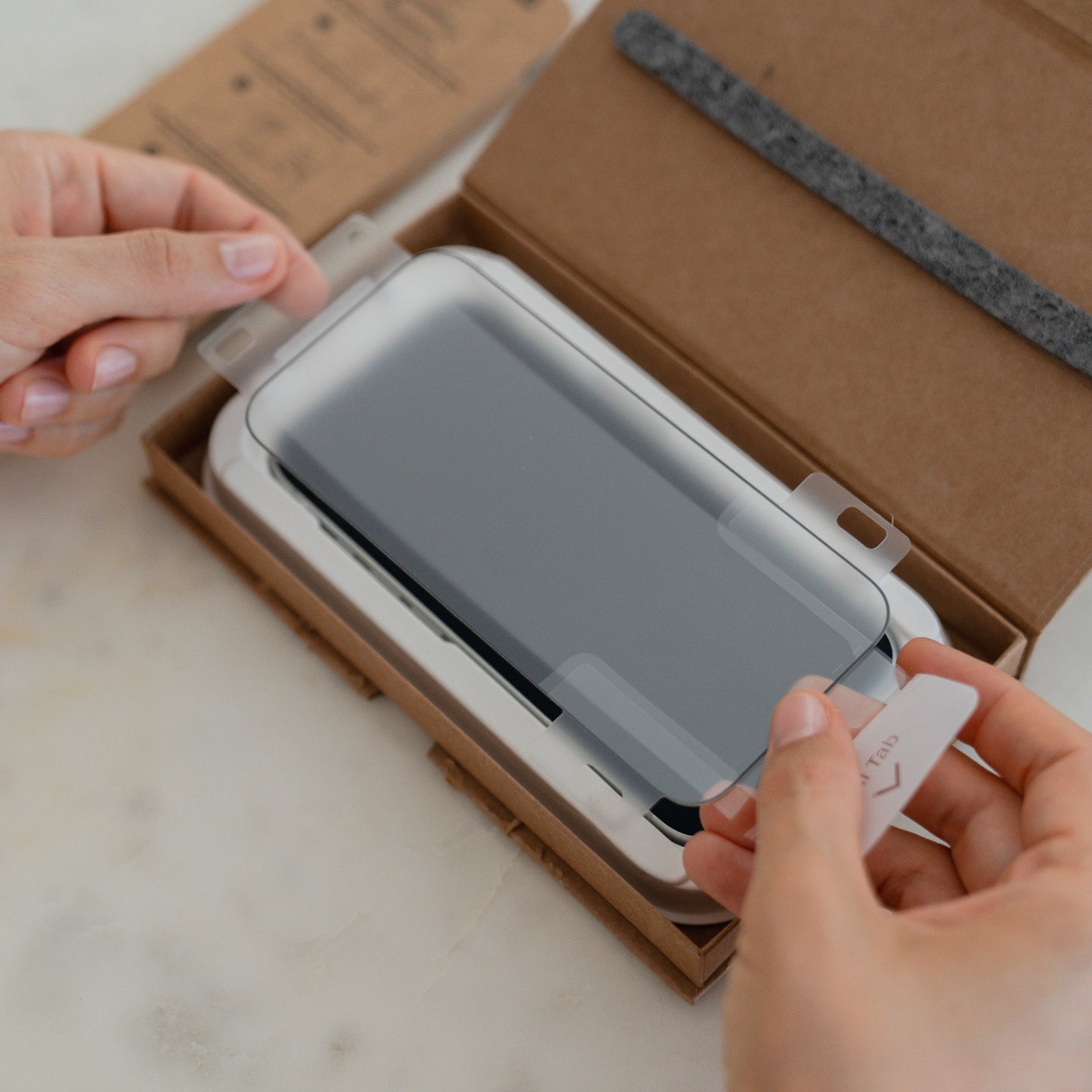 Hände, die einen arktis 2er Set iPhone 17 Pro Max REVOLUTION Glas-Displayschutz auf ein Smartphone aufbringen, entnommen aus einer braunen arktis-Kartonverpackung.
