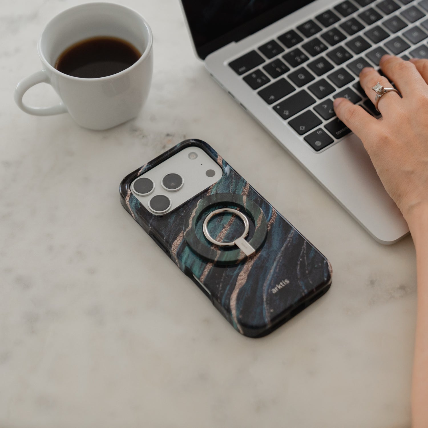 Eine Hand ruht auf einem Laptop, einer Kaffeetasse und einem Smartphone, während der „Arktis Magnetischer Ringhalter für MagSafe – Green Marble“ – ein unverzichtbares iPhone-Zubehör – auf einem Marmortisch liegt.