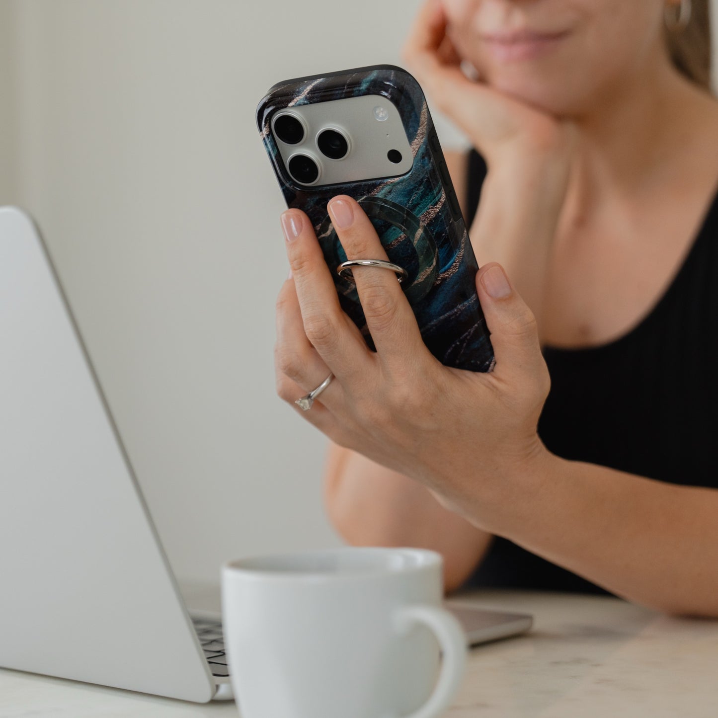 Eine Frau sitzt an einem Tisch neben einem Laptop und einer Kaffeetasse, lächelt leicht und hält dabei ihr Smartphone in der Hand, das mit dem „Arktis Magnetischer Ringhalter für MagSafe – Green Marble“ ausgestattet ist.