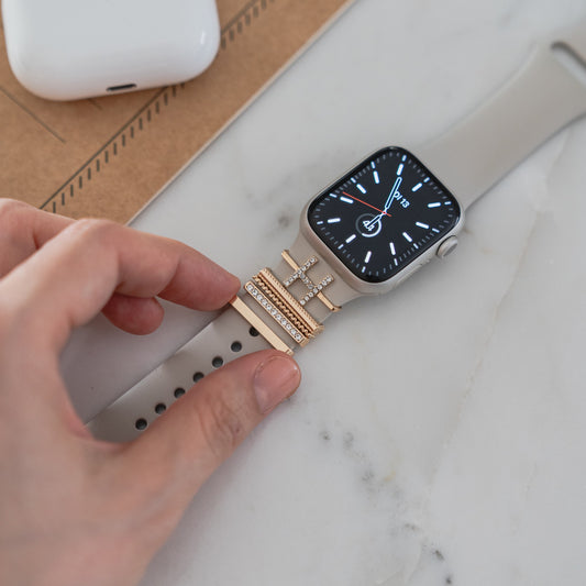Eine Hand befestigt die arktisband Apple Watch Charms „H Style“ von arktisband an einem beigen Smartwatch-Armband auf einer Marmoroberfläche.