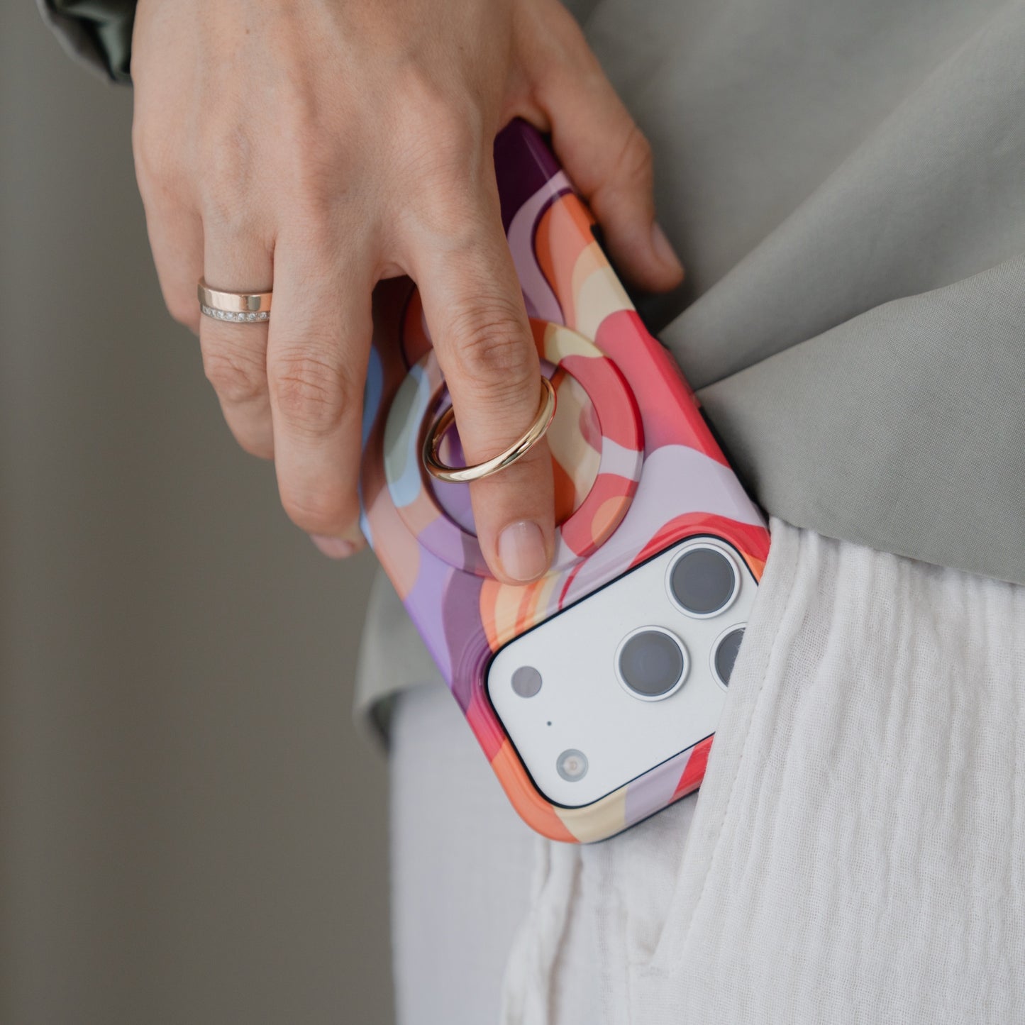 Eine Hand, die ein farbenfrohes Smartphone mit dem „Arktis Magnetischer Ringhalter für MagSafe – Happy Life“ hält und es in die Hosentasche einer weißen Hose steckt – ein ideales iPhone-Zubehör für stilvollen Komfort.