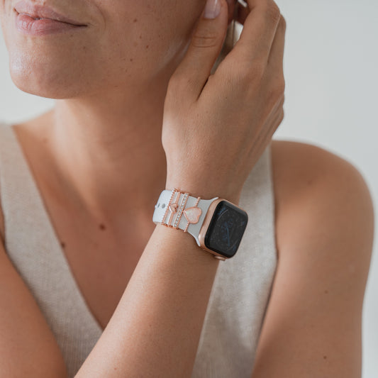 Eine Person in einem ärmellosen hellbeigen Oberteil berührt ihr Ohr und präsentiert das arktisband Apple Watch Charms „Loyal Roségold“ Accessoire von arktisband an einem weißen Armband mit zwei roségoldenen Armbändern. Das Display der Uhr ist dunkel und nur teilweise sichtbar.