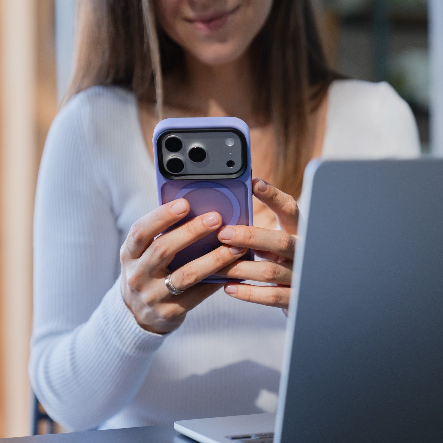Eine Frau hält ein lilafarbenes Arktis iPhone 17 Pro Solid Case mit MagSafe - ein mattes, MagSafe-kompatibles Case von Arktis - auf ihr Smartphone, während sie vor einem offenen Laptop sitzt.