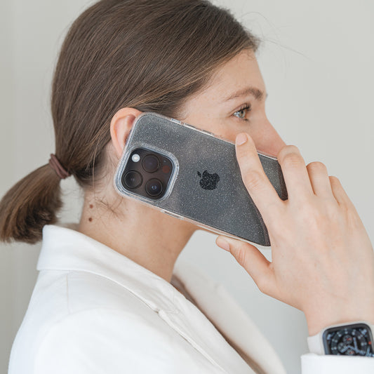Eine Frau in einem weißen Blazer mit zurückgebundenen Haaren hält ein iPhone 14 Pro Max in einem Blingmeister CRYSTAL GLITTER Case an ihr Ohr und trägt eine Smartwatch.