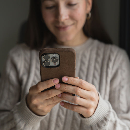 Eine Person in einem beigen Zopfmusterpullover hält eine arktis iPhone 15 Hülle aus Alcantara in Braun und lächelt auf den Bildschirm. Der Fokus liegt auf ihren Händen und dem Telefon; der Hintergrund ist unscharf.