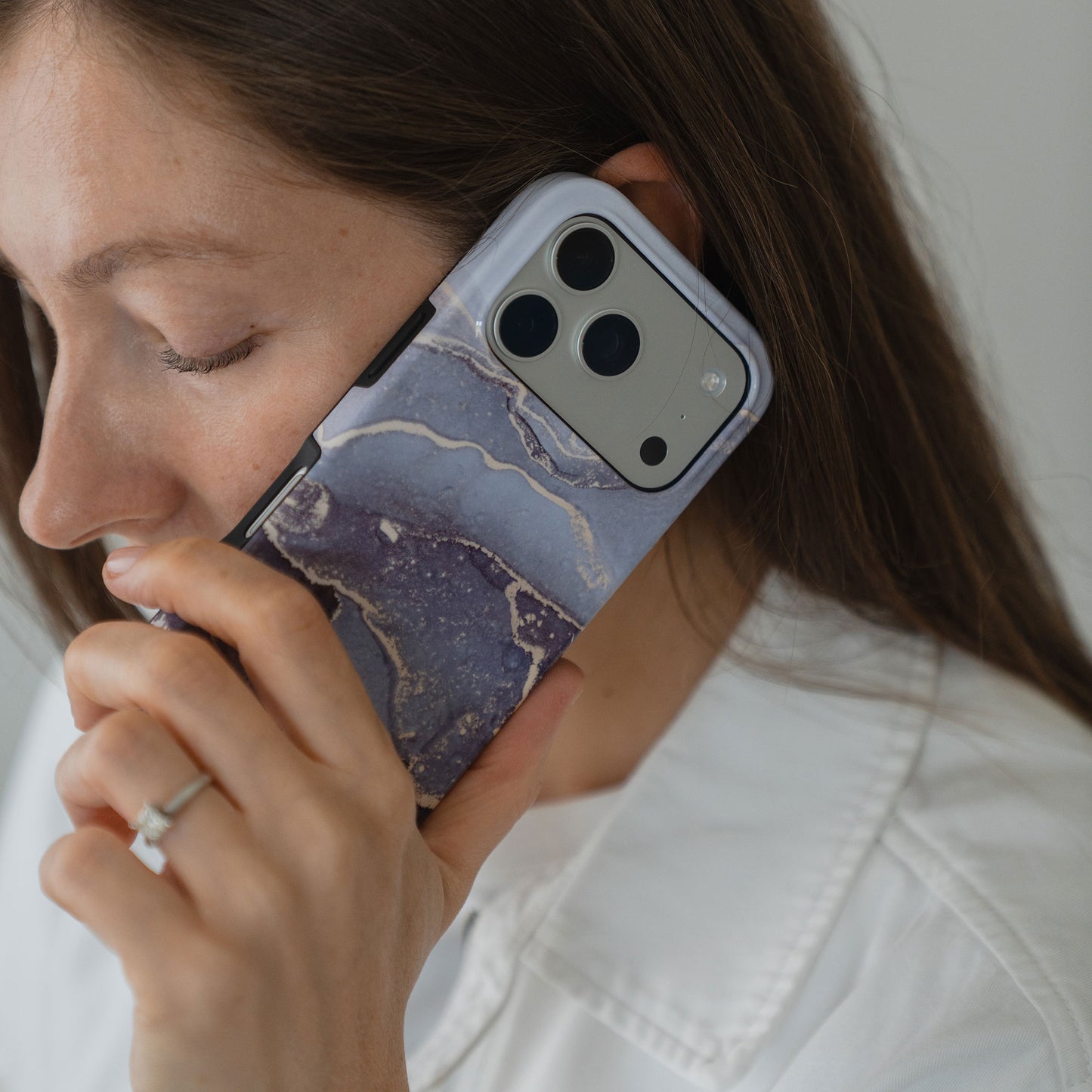 Eine Frau mit weißem Hemd und Ring hält eine arktis iPhone 14 Pro Max Hülle - Dusty Marble an ihr Ohr. Die MagSafe-kompatible Hülle von arktis verfügt über stoßabsorbierendes TPU-Silikon in einem lila Marmor-Design.