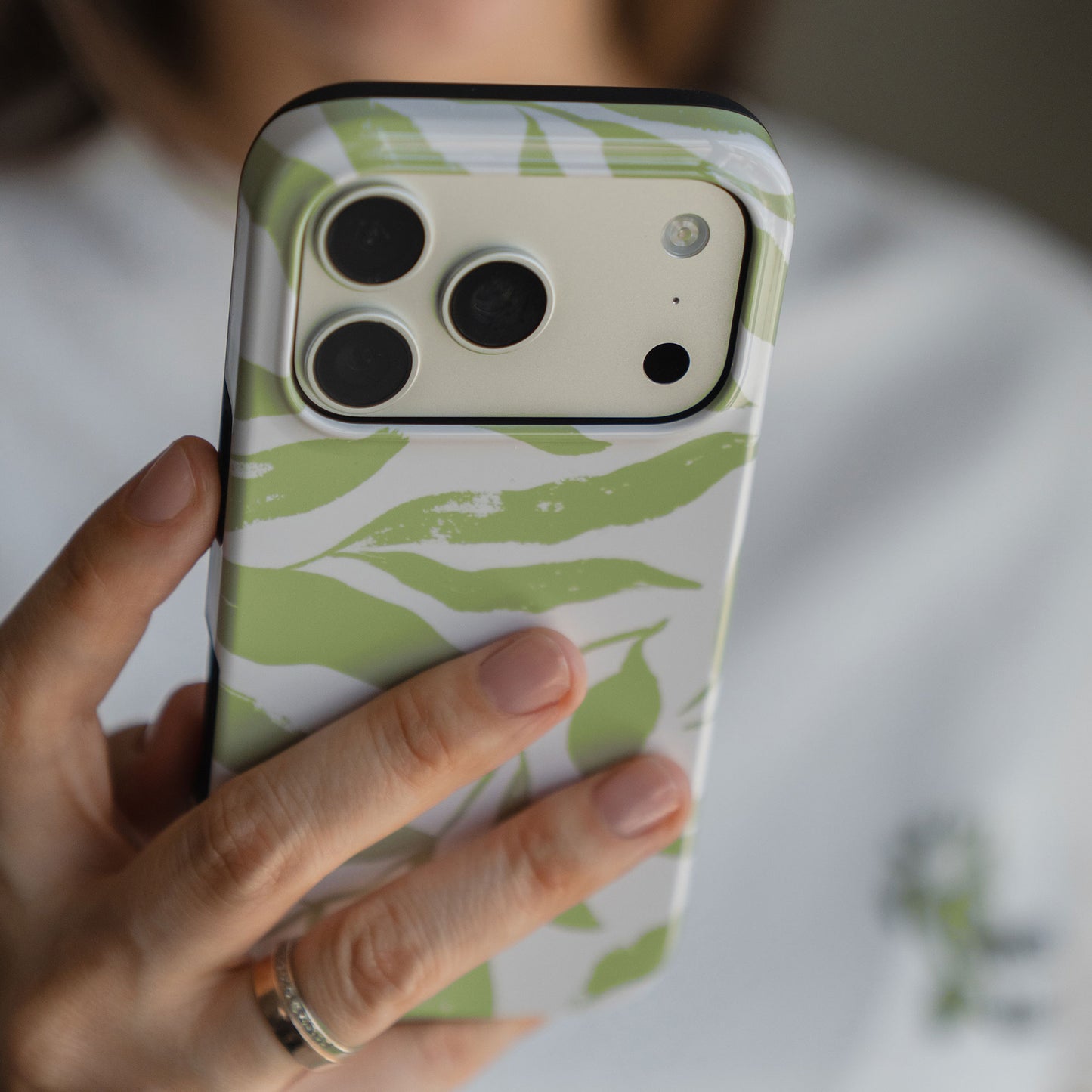 Eine Hand hält die arktis iPhone 14 Pro Max Hülle - Botanic, eine MagSafe kompatibele Hülle mit grünem und weißem Blätterdesign von arktis, ausgestattet mit stoßabsorbierendem TPU-Silikon für zusätzlichen Schutz der Kameralinse.