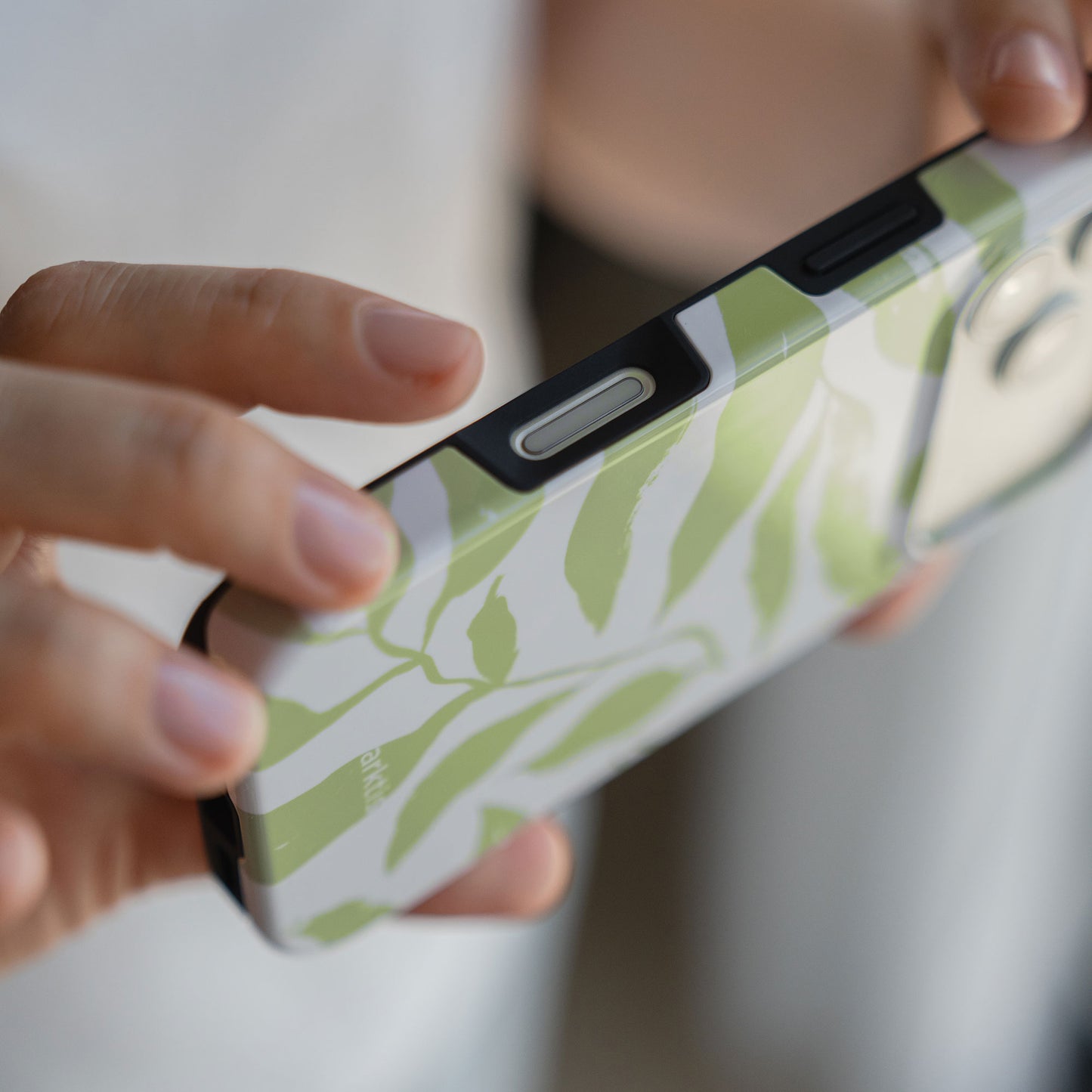 Eine Person hält eine arktis iPhone 14 Pro Max Hülle - Botanic, wobei die seitlichen Tasten hervorgehoben werden. Die grün-weiß blattgemusterte MagSafe-kompatible Hülle von arktis bietet zusätzlichen Schutz mit stoßabsorbierendem TPU-Silikon.