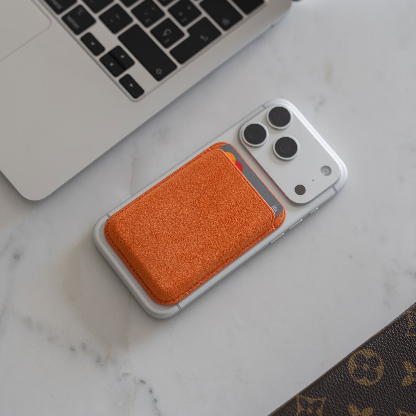 Ein iPhone mit dem arktis iPhone MagSafe Kartenhalter aus Alcantara liegt auf einem weißen Schreibtisch neben einem Laptop.