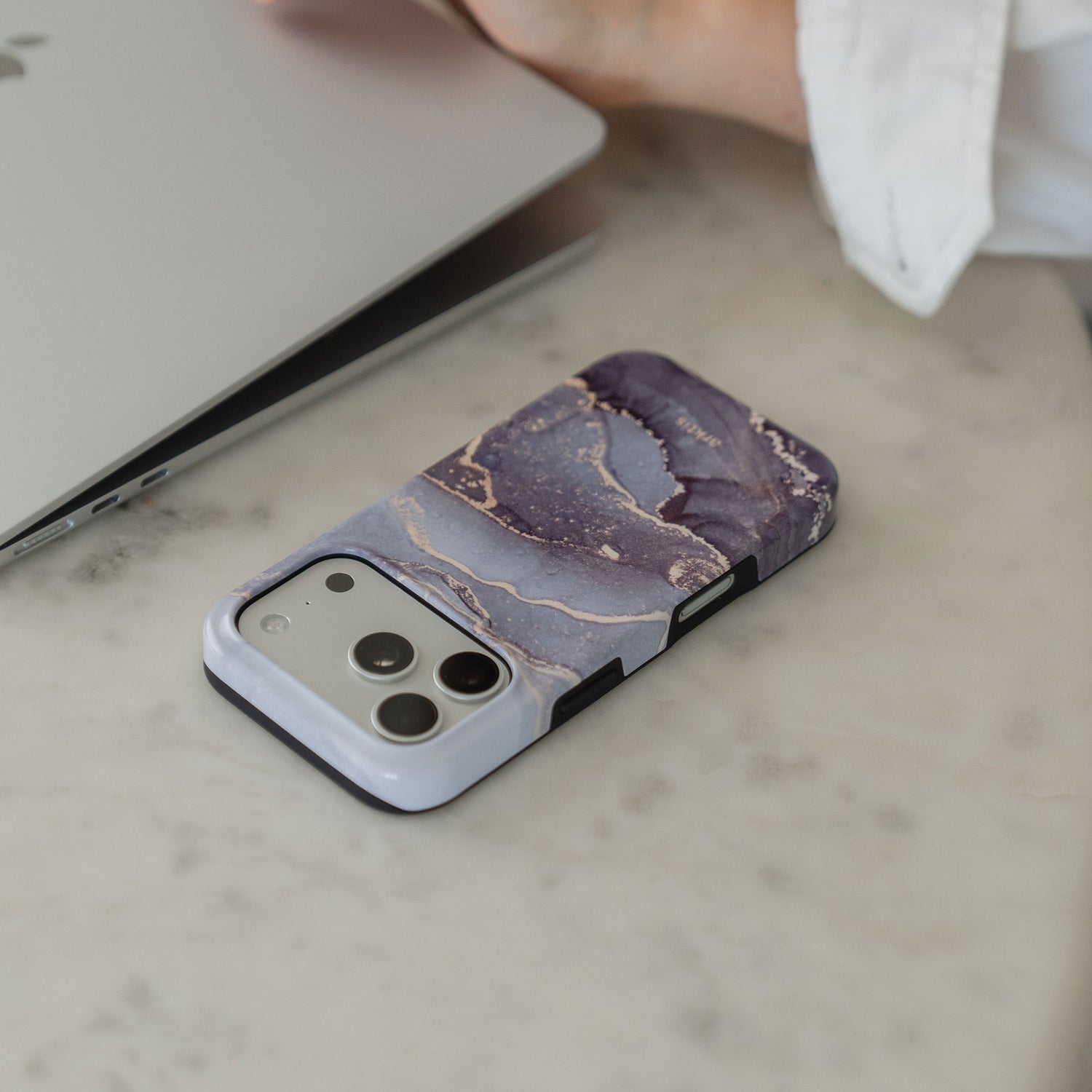 Die arktis iPhone 14 Pro Max Hülle - Dusty Marble von arktis, eine MagSafe kompatible stoßabsorbierende TPU-Silikonhülle im lila Marmor-Look, liegt auf Marmor neben einer Hand und einem Laptop.