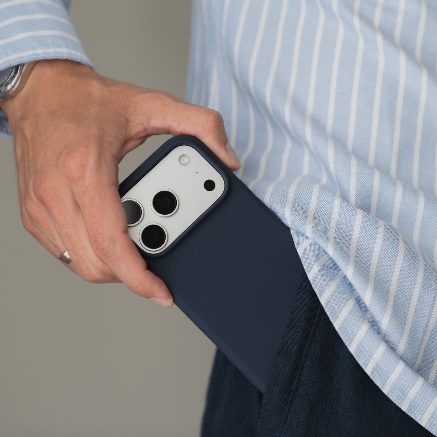 Hand mit einem arktis iPhone 18 Pro Max Silikon-Case mit MagSafe in der Farbe Mitternacht; das Handy wird in die Tasche gesteckt; die Person trägt ein gestreiftes Hemd.