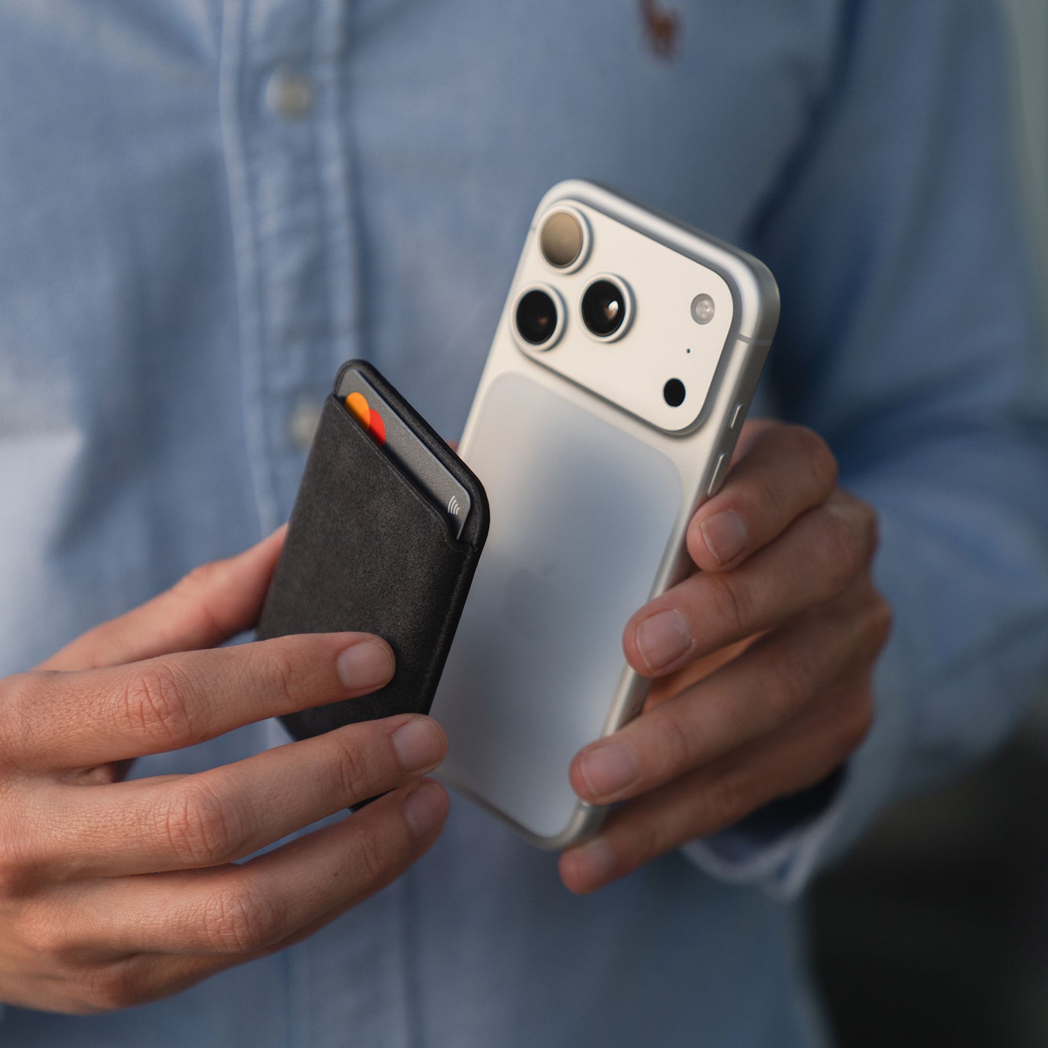 Person, die ein weißes Smartphone mit dem arktis iPhone MagSafe Kartenhalter aus Alcantara wallet by arktis und einer schlanken schwarzen Alcantara-Brieftasche mit einer sichtbaren Karte in der Hand hält, trägt ein blaues Button-up-Hemd.