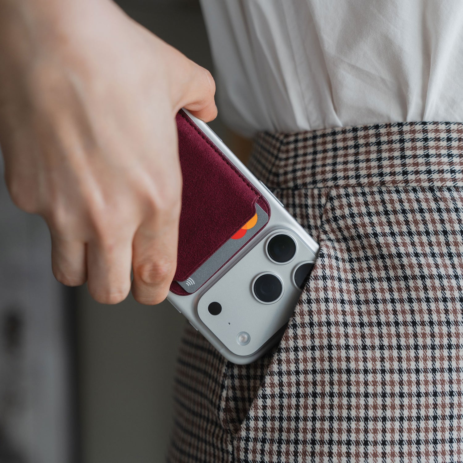 Eine Person steckt ihr iPhone, ausgestattet mit einem arktis iPhone MagSafe Kartenhalter aus Alcantara, in die Tasche einer karierten Hose.