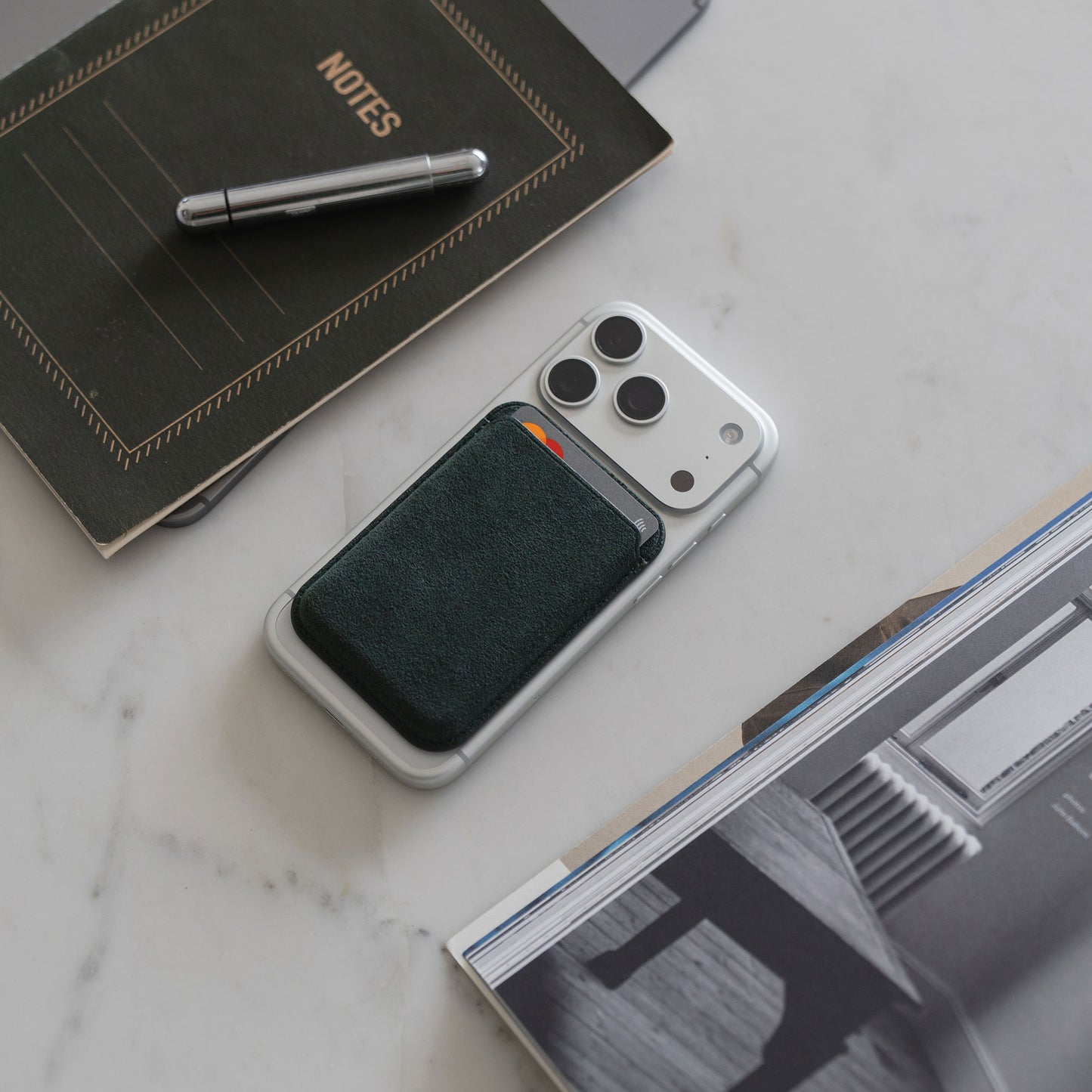 Ein arktis iPhone MagSafe Kartenhalter aus Alcantara, iPhone 12, Stift, Notizbuch und Zeitschrift sind auf einer Marmorfläche angeordnet.