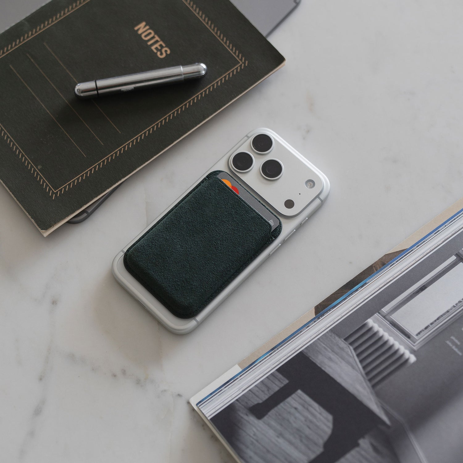 Ein arktis iPhone MagSafe Kartenhalter aus Alcantara, iPhone 12, Stift, Notizbuch und Zeitschrift sind auf einer Marmorfläche angeordnet.