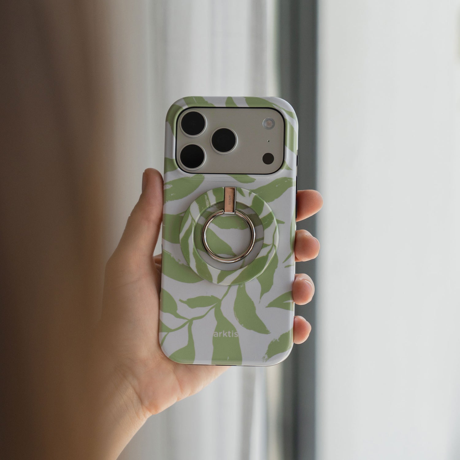 Eine Hand hält die arktis iPhone 14 Pro Max Hülle - Botanic von arktis, eine grüne, blattgemusterte Hülle mit kreisförmigem Ringhalter aus stoßabsorbierendem TPU-Silikon.