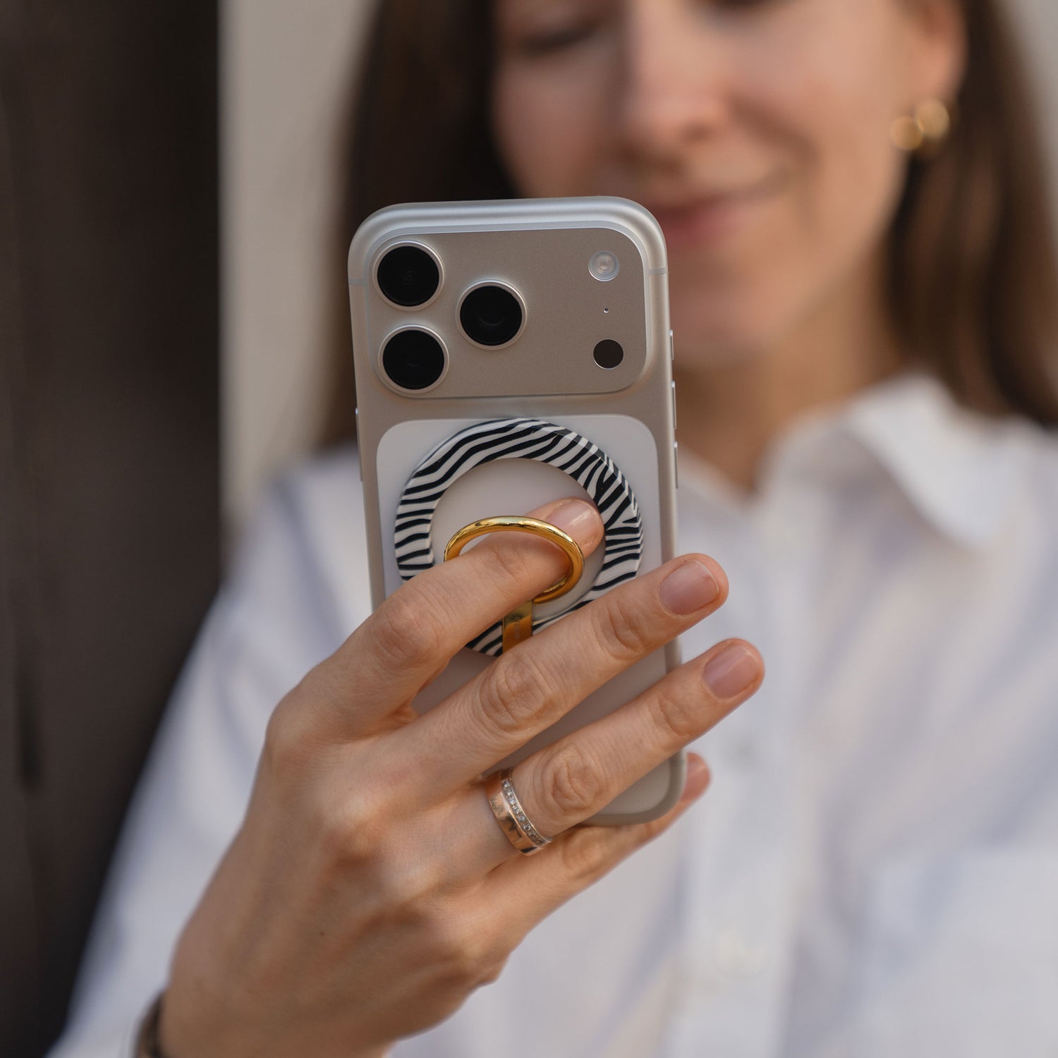 Eine Frau hält ihr Smartphone mit dem arktis Magnetischer Ringhalter für MagSafe - Sweet Leopard. Sie trägt eine weiße Bluse und goldene Ohrringe - stylisches iPhone Zubehör mit starken N52 Magneten für sicheren Halt und trendigem Leopardendesign.