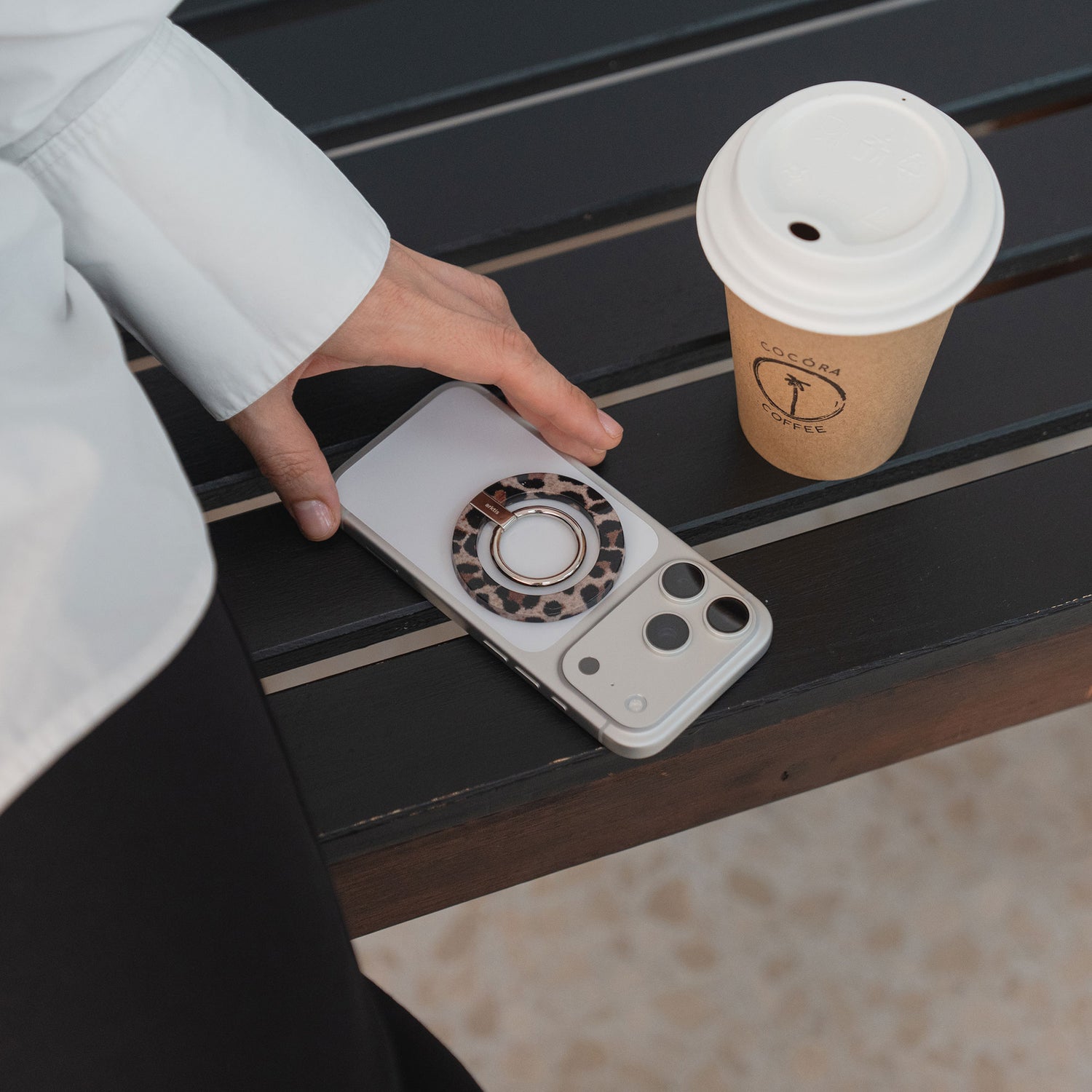 Eine Hand greift nach einem Smartphone neben dem arktis Magnetischer Ringhalter für MagSafe - Sweet Leopard neben einem Pappbecher auf einer dunklen Holzbank.