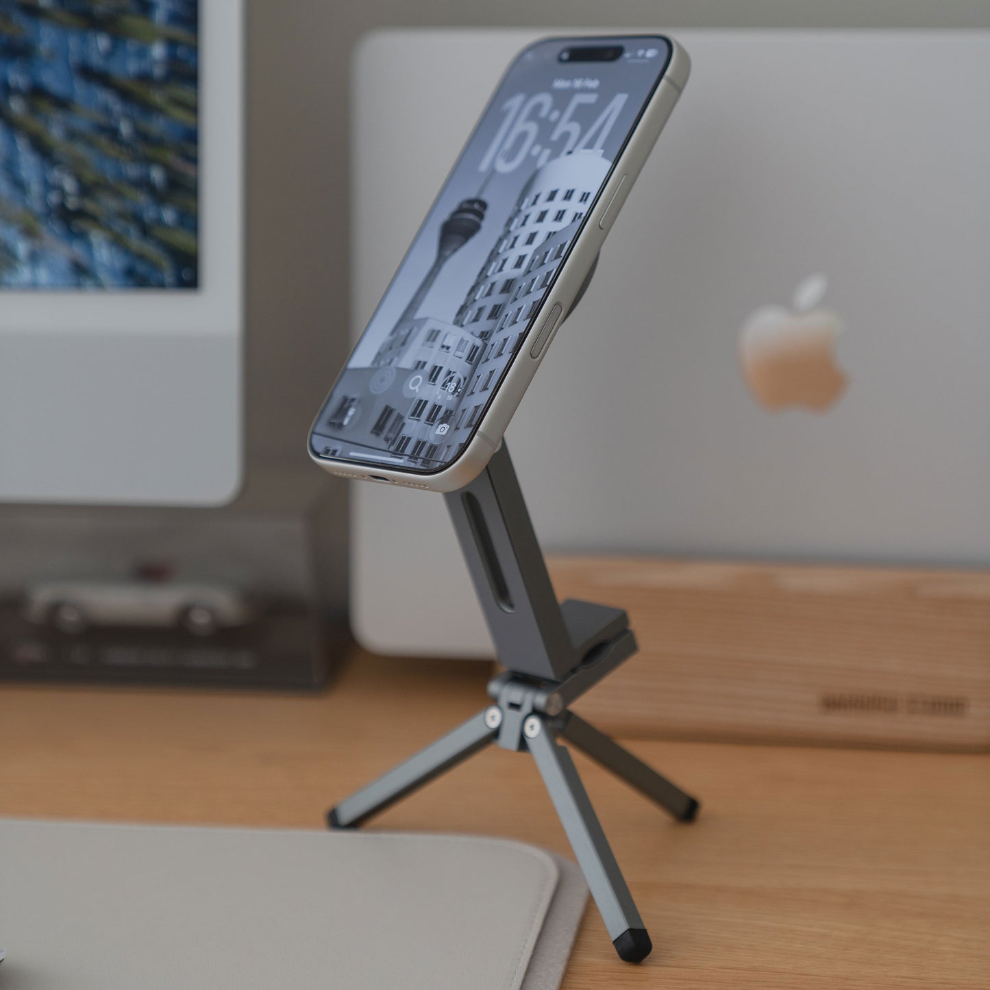 Ein iPhone mit der arktis iPhone MagSafe Tripod 360 Halterung von arktis wird auf einem Holztisch vor einem iMac Computer platziert.