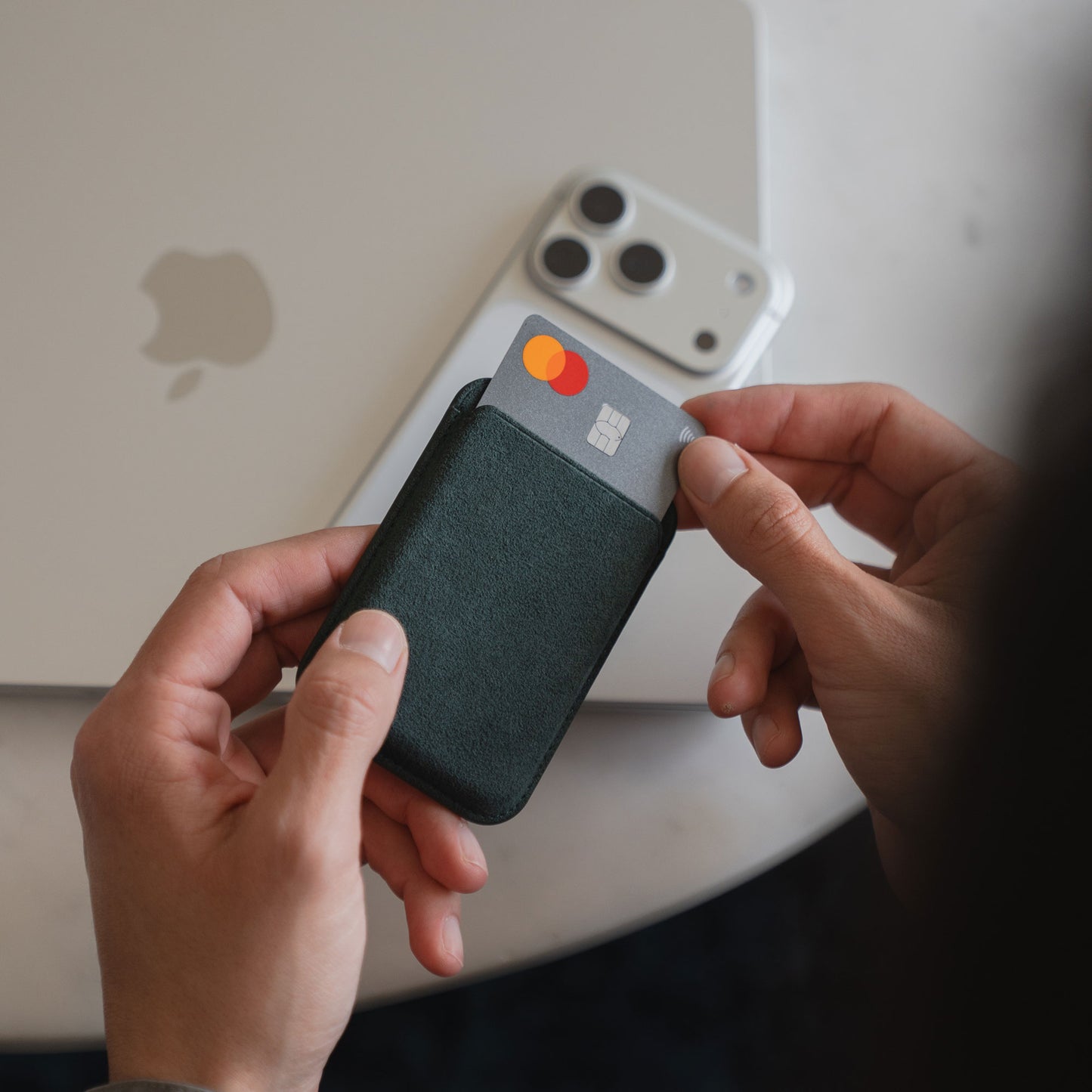 Die Hände halten ein Portemonnaie mit einer Kreditkarte neben einem weißen iPhone 12, dem arktis iPhone MagSafe Kartenhalter aus Alcantara von arktis, und einem geschlossenen Laptop auf dem Tisch.