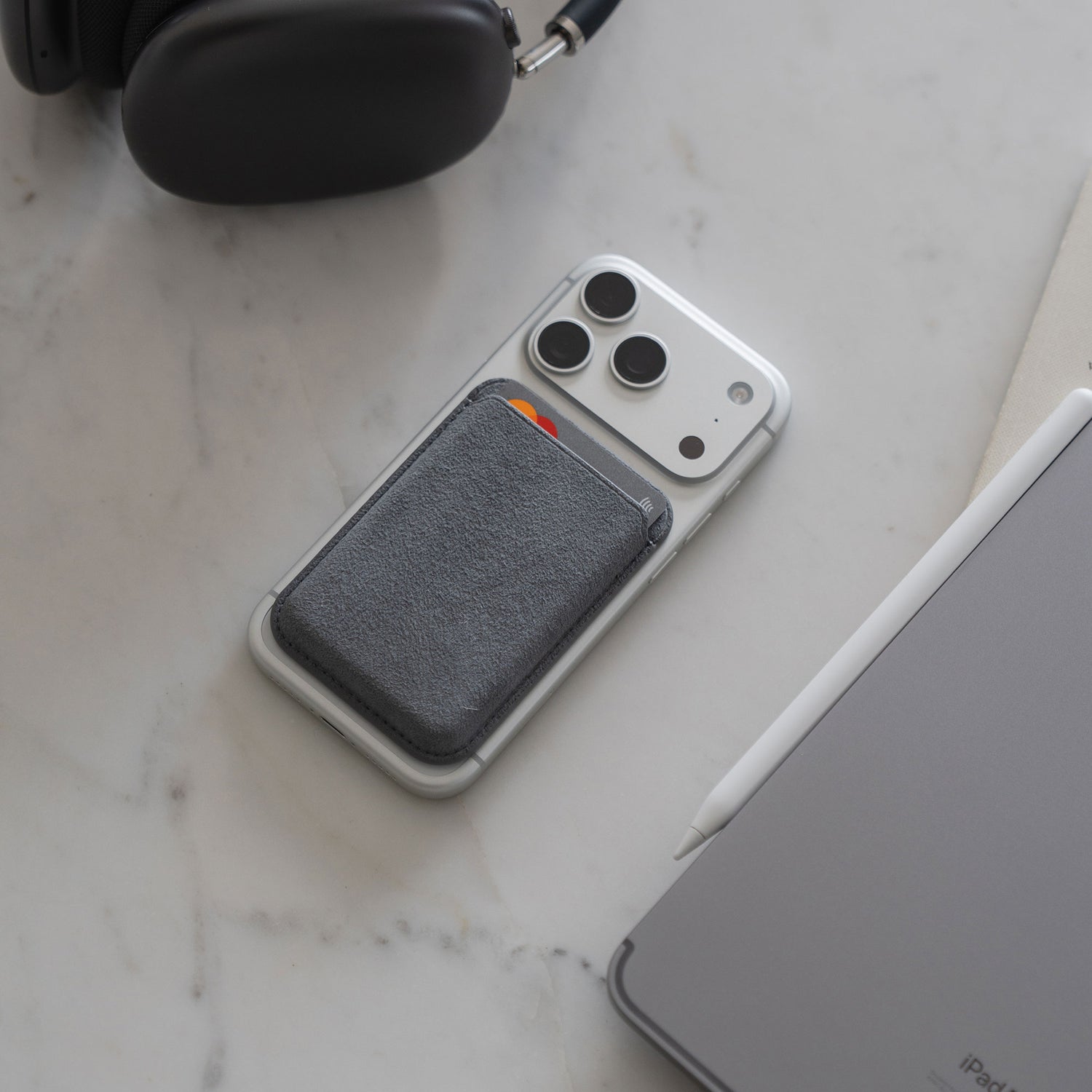 Ein arktis iPhone MagSafe Kartenhalter aus Alcantara ist an einem iPhone befestigt, das neben Kopfhörern, einem Tablet und einem Stylus auf einer Marmorfläche liegt.