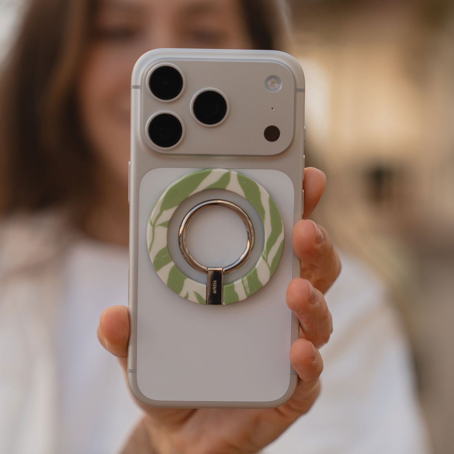 Eine Person hält ein Telefon mit dem arktis Magnetischer Ringhalter für MagSafe - Botanic in grün und weiß, der stilvolles iPhone Zubehör bietet, das durch starke N52-Magnete gesichert wird.