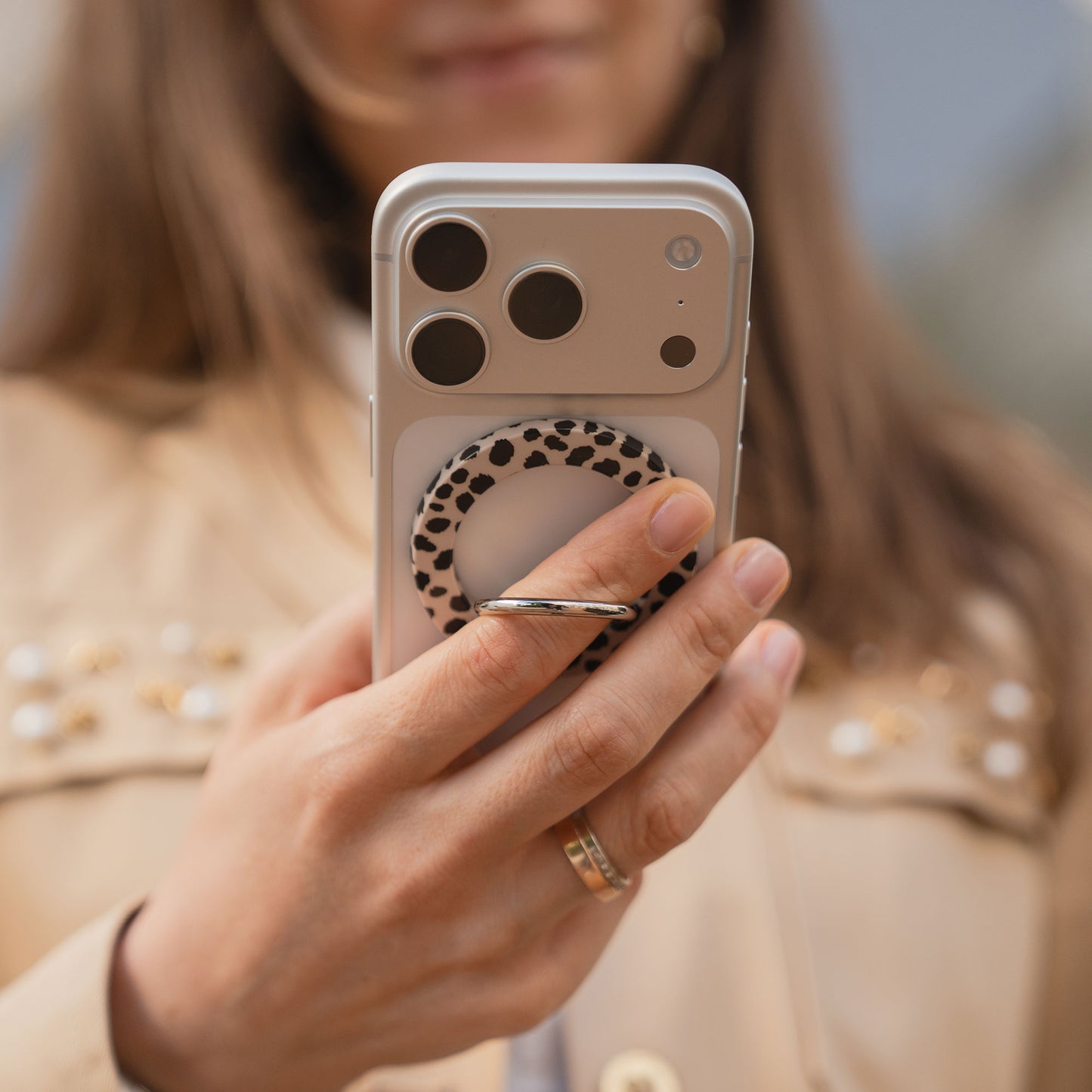 Eine Person hält ihr Smartphone mit dem arktis Magnetischer Ringhalter für MagSafe - Sweet Leopard, mit N52 Magnete, während sie eine beige Jacke und einen goldenen Ring trägt - das ideale stylische und funktionale iPhone Zubehör.