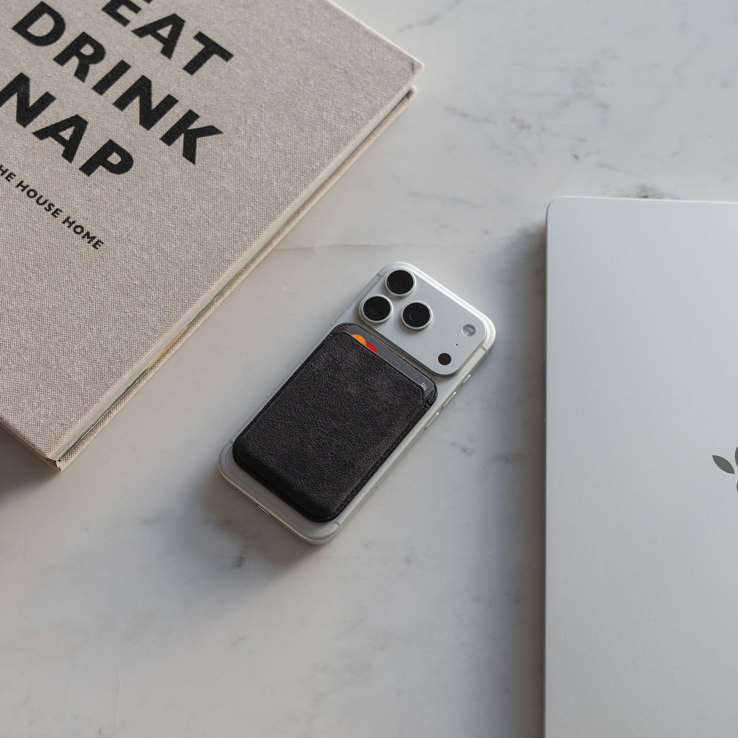 Ein iPhone mit dem arktis iPhone MagSafe Kartenhalter aus Alcantara von arktis ruht auf einem weißen Schreibtisch neben einem Laptop und einem Buch.
