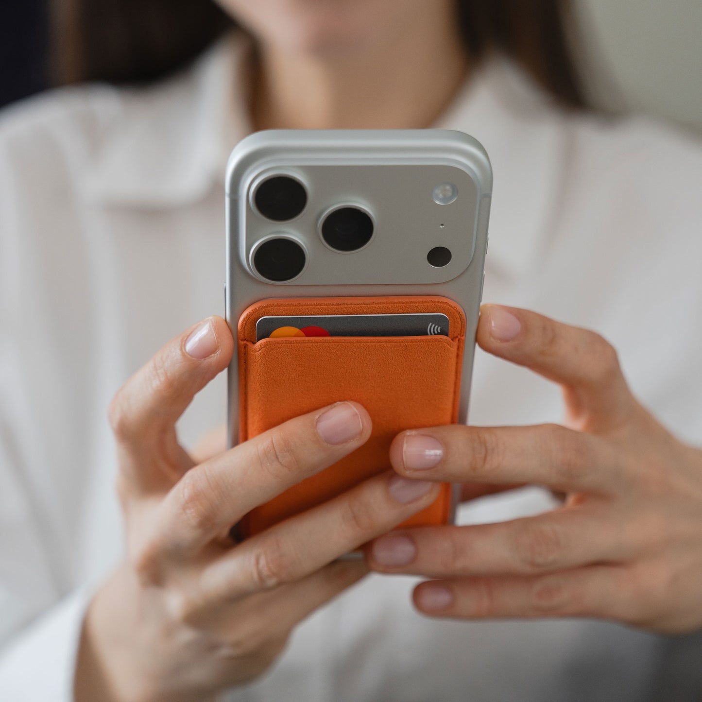 Eine Person hält ein iPhone mit dem arktis iPhone MagSafe Kartenhalter aus Alcantara von arktis in leuchtendem Orange, wobei eine Kreditkarte teilweise im Halter sichtbar ist.