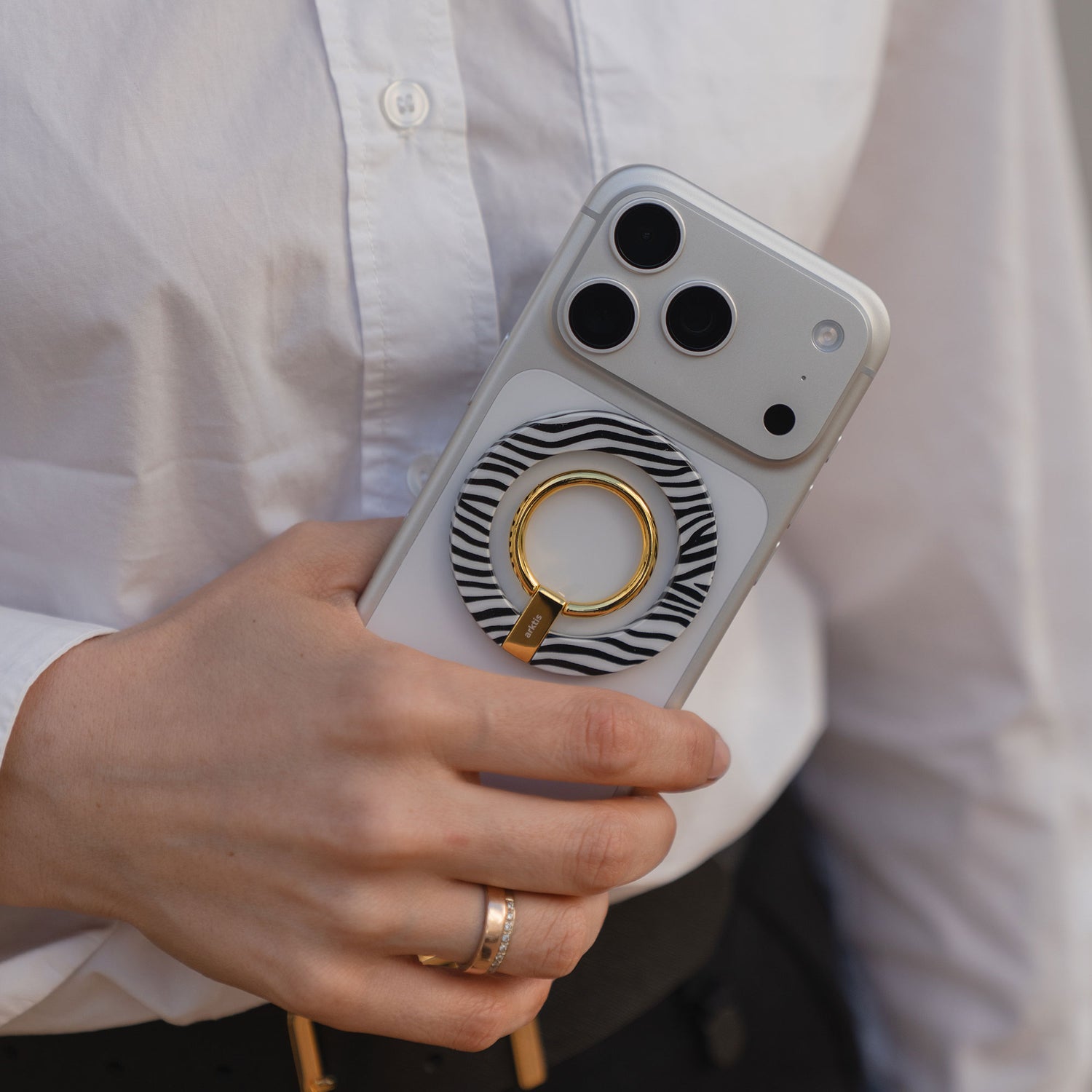 Eine Person hält ein iPhone mit dem arktis Magnetischer Ringhalter für MagSafe - Sweet Leopard und kombiniert ihn mit einem weißen Hemd und goldenen Ringen für eine schicke Mischung aus Stil und funktionellem Zubehör.