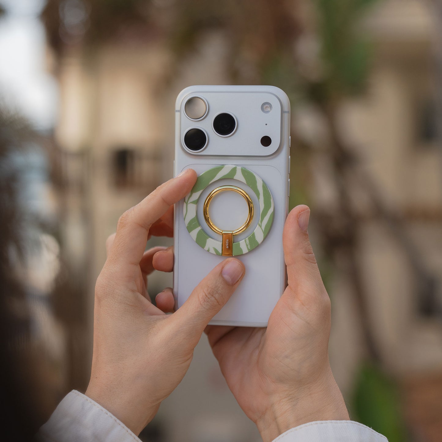Eine Person hält ein weißes Smartphone, auf dessen Rückseite der arktis Magnetischer Ringhalter für MagSafe - Botanic in Grün und Gold angebracht ist, der für stylisches iPhone Zubehör sorgt.