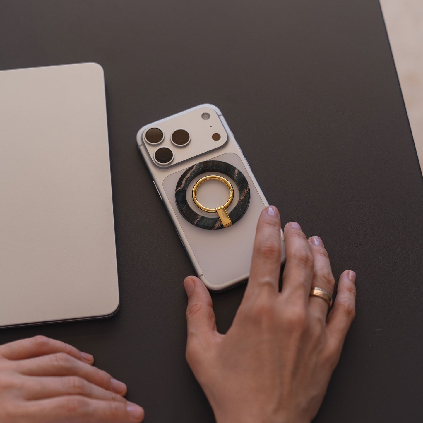 Eine Hand mit einem Goldring ruht neben einem arktis Magnetischer Ringhalter für MagSafe - Sweet Leopard, abgebildet mit iPhone Zubehör und einem geschlossenen Laptop auf einer dunklen Fläche.